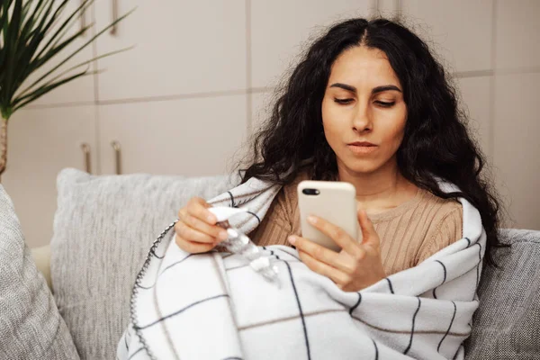 stock image Beautiful young arab woman sitting on the couch in her room wrapped in a blanket. Holds a smartphone in one hand and an electronic thermometer in the other pill. She dials the family doctor's phone.