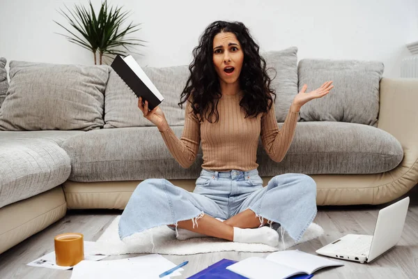 Beautiful young Spanish woman sitting on the floor in her house. Girl student faced a problem in studying and she spreads her hands. Student overwhelmed with paperwork.