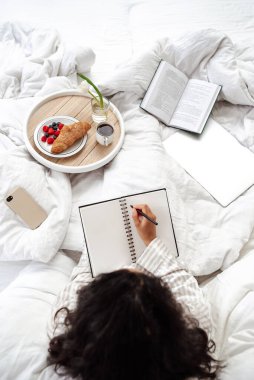 Yatakta rahat bir rahatlama. Beyaz pijamalı bir kadın yatakta kahvaltıyla not alıyor. Bir kitap okuduktan sonra hayal kuran ve gelecek için notlar alan bir kadın.