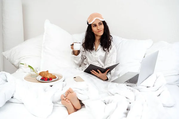Güzel beyaz blog yazarı dizüstü bilgisayar teknolojisiyle otel odasındaki dinlenme günü spasında gülümsüyor dijital netbook teknolojisi olan neşeli bir kadın serbest yazar oda servisinden keyif alıyor.