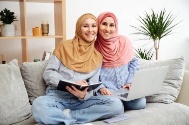 Doğulu genç öğrenciler evde tesettüre hazırlanıyorlar. Bir dizüstü bilgisayar ve dizüstü bilgisayarla kanepede oturup not defterine, klasöre kalemle yazmak. Kameraya gülümsüyor..