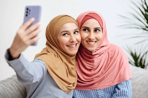 İki güzel Müslüman kadın video kaydediyor, parlak bir şekilde gülümsüyor, selfie çekiyor, yeni bir bakış açısı yakalıyor, pembe atkı ve kazakla poz veriyor, takipçiler için internete fotoğraflar yüklüyor, kapalı mekanda güzel fotoğraflar çekiyor.