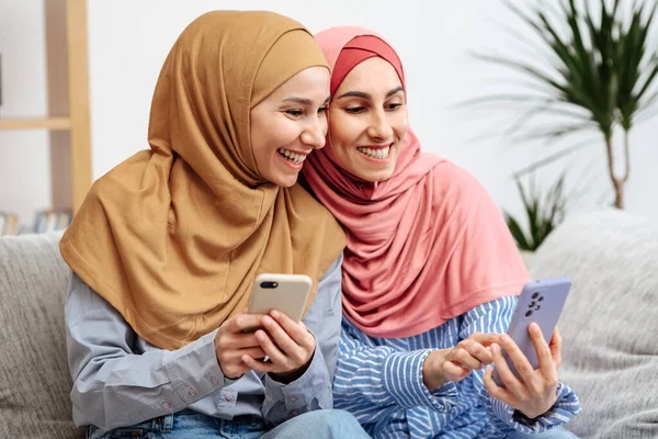 Mutlu iki Arap kadın akıllı telefondan iyi haberler okuyor, kızlar evde kanepede online içeriği izlemek için telefon kullanıyor. Kadınlar başörtüsü takar ve günlük kıyafetler giyerler..