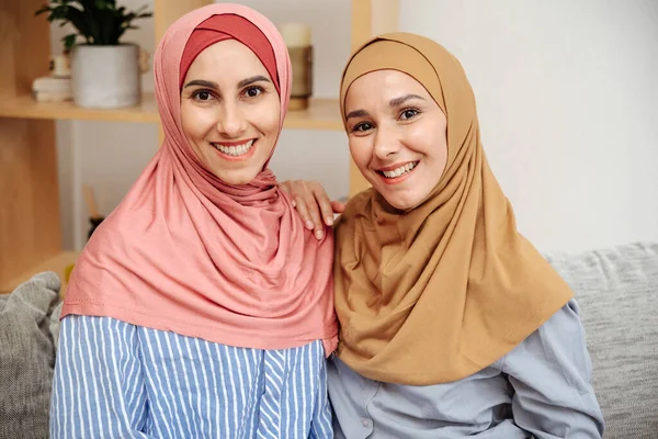 Portrait of two muslim women in hijabs sitting on sofa at home. Girls smile pleasantly and enjoy the weekend together. Two Arab in casual clothes. Bright smiling faces looking directly at the camera