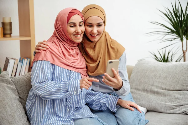 Çevrimiçi bankacılık. tesettürlü müslüman kadınlar evdeki akıllı telefonda uygulama kullanıyor oturma odasında koltukta oturuyor, uzayı kopyalıyor. Kızlar sarılıp telefon ekranına ve daktiloya bakar.