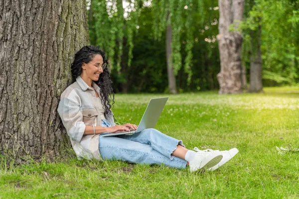 E-learning Concept. Arab Female Student With Laptop Sitting Under Tree In Park, Smiling Young Middle Eastern Woman Study Online On Computer While Relaxing On Green Lawn Outdoors, Copy Space