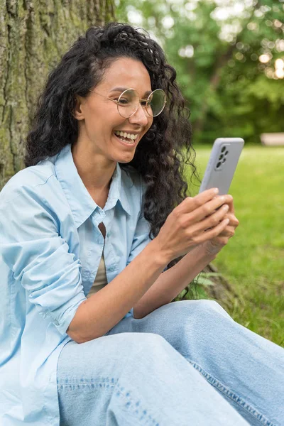 Güzel İspanyol kadın arkadaşlarıyla mesajlaşır ve mesajlaşır. Telefonda gördüklerine sevinir. Mavi günlük kıyafetlerle bir telefon tutar, güler, şehir parkındaki açık havaya bakar.