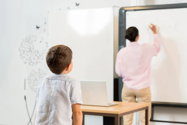 生徒と教師のリアビュー 教師はブラックボードのタスクを書き 子供は好奇心を持って見ています 小学生トレーニングのコンセプトと長期休暇の後に戻る — ストック写真