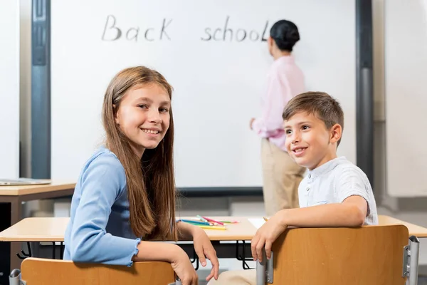 学習コンセプト 前景には 机に座っている2人の子供が振り向いてカメラを見ています 女性教師が黒板に書いている 子供たちが学校の年を始める — ストック写真