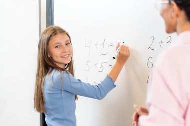 Avrupa görünümlü mutlu öğrenci tahtaya matematiksel bir örnek yazıyor. Görevleri kolayca çözen ve kameraya bakan bir kızın portresi. Yakınlarda bir öğretmen duruyor ve yardım ediyor..