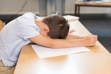 Tired bored schoolboy, primary student in online school, sitting at desk, leaning on the table with his forehead, having difficulty adapting to the new academic year semester. Homeschooling