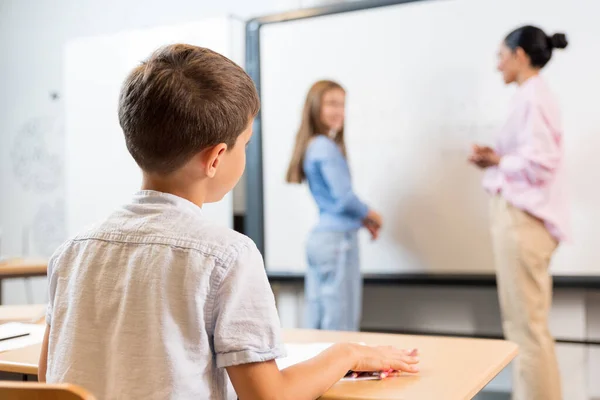 机の上に座っている教室の小さな男の子のリアビュー ボーイはブラックボードの近くで答える先生と彼の同級生を見ています 少年は自分の答えを心配している — ストック写真