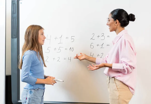 教室で数学を解決しながら 教師に質問する女子高生の側面 混血した女性教師が女の子をタスクで助けます 研究テーマについての活発な会話 — ストック写真