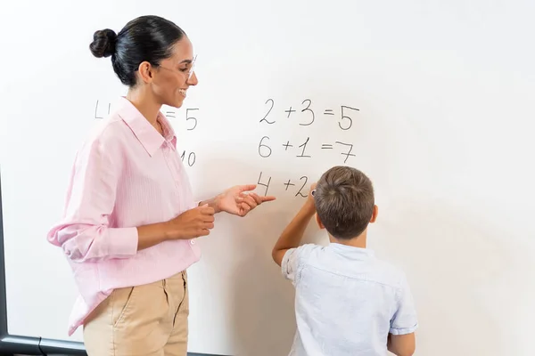 ブラックボードの近くに立って数学の仕事を解決する 混合されたレースの女性教師と小さな学校のボーイを笑顔で 現代のライト教室での学習と解決の概念 — ストック写真