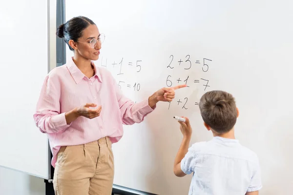 ハッピースクール教師が黒板に追加する学校のボーイを手伝ってくれました メガネの女性教師は 数学の例を学校のボーイに説明しています 学習に関するコミュニケーションとディスカッション — ストック写真