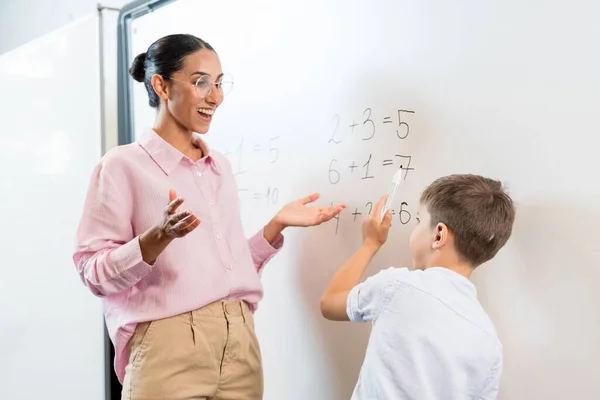 ホワイトボードに小さなボーイが書いている中東の教師の笑顔を閉じます 先生は彼女の生徒の成功に驚いています 少年は数学的な例をうまく解決しました — ストック写真