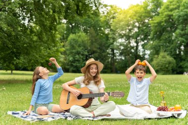 Hasır şapkalı tatlı sarışın kız şehir parkında çimlerin üzerinde gitar çalıyor. Küçük erkek ve kız kardeş battaniyeye oturup yemekle oynayın, eğlenin. Piknikte üç çocuk.