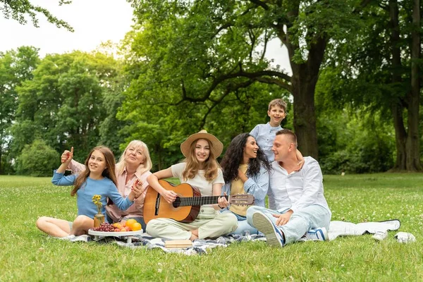 Picnic, big family resting on the grass for summer vacation, outdoor recreation and healthy lifestyle together. Grandparents, mother and children with a guitar in a field park