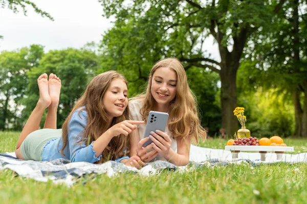 İki tatlı sarışın kız parkta çimlerin üzerinde battaniyeye uzanmış, telefonu ellerinde tutuyorlar, haberleri izliyorlar. Piknikte sohbet eden kızlar, telefon ekranına dokunmak, kaydırmak ve kaydırmak