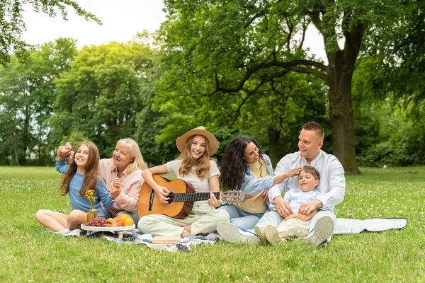 Cheerful friendly family with different generations of mixed race is sitting in the city park on a picnic. Play songs on the guitar, listen and sing, fun and hug. Concept of family outdoor recreation.