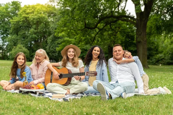 Happy multiracial family relaxing together. Diverse young people sitting on grass in city park. Happy hour, lunch break and youth concept. Teenager in a hat plays the guitar, everyone smiles.