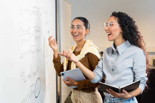 2人の美しいヒスパニックの女性学生が教室の黒板の近くに立って黒板を指し 公式の正しい解決策に喜んでいます 女の子は大学の試験にうまく準備されています — ストック写真