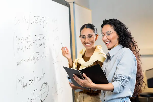 黒板の近くの教室に立っている2人の美しい女性の外観の学生は 数学の方程式を解くノートブックの笑顔を見て 正しい解決策を見ます チームワークが成功につながる — ストック写真