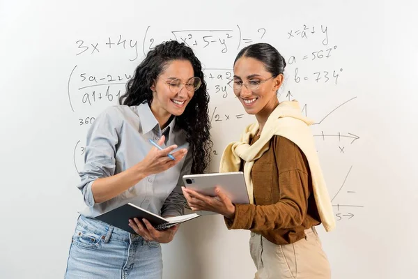 2人のかわいいアラブの女性学生が黒板の近くに立って タブレットの計算を見ます 笑顔でカジュアルな服を着たプロジェクトについて話し合う女性たち 共同学習コンセプト — ストック写真