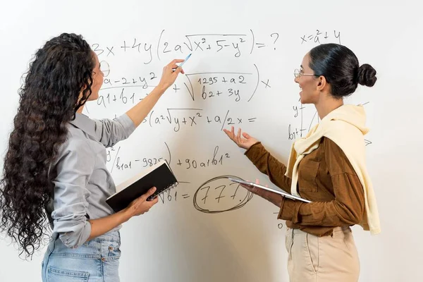 中東の2人の女性学生が黒板の近くに立って方程式を示しています 女性はタブレットとタブレットを手に持っているタスクを共同で解決します カールヘアの女の子は教育を行います — ストック写真