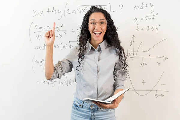 数学のタスクを持つ黒板の背景に立っている眼鏡でかわいいカーリー髪のアラブ女性の肖像画 女性にはアイデアがあります 少女はノートを保持し 指を上げました — ストック写真