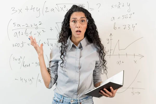 ストレス バーンアウト 眼鏡のカーリーヘアの女性教師が手を振り 驚いたことにカメラを見つめている 背景は数学的なグラフと方程式を持つブラックボードです — ストック写真