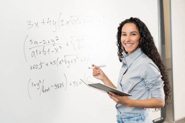 Gömlek giyen ve sınıfta kameraya bakan gülümseyen Doğulu iş kadını. Kıvırcık Arap öğretmen ya da profesör beyaz tahtanın önünde portre için poz veriyor. Bir dizüstü bilgisayar, işaretleyici