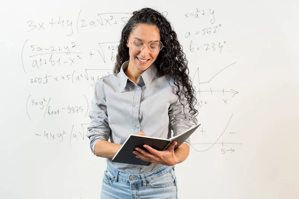 年轻的西班牙裔女学校数学老师 大学导师 教练看着笔记本 边写边笑 背景是一个有数学任务的白板 教育和课程管理的概念 — 图库照片