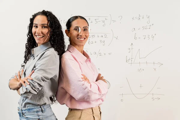 两个高中生抱着胳膊站在教室里笑着 穿着商务服装戴着东方风貌眼镜的布鲁内特女孩 背景是一块有数学任务的黑板 — 图库照片