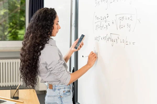 東洋の外観の美しい若い教師は レッスンに備えているタスクを書き留めています 教室でブラックボードを書いている間 スマートフォンを使用して過激な計算をする女性 サイドビュー — ストック写真