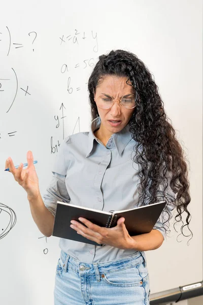 戴眼镜的卷发阿拉伯女学生站在黑板旁边 看着笔记本 不知道如何解决这个问题 在计算和计算中弄混 — 图库照片