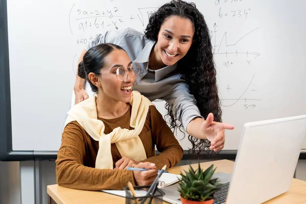 中東の女性の若い友好的なビジネスはお互いに話し合い 現代の教室で机に座ってラップトップに油を注いでいます 背景にはブラックボードがあります — ストック写真