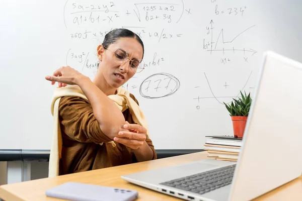 Distant education, remote job and health concept - tired Mixed Race female teacher with laptop computer touching her elbow at office. Worried woman feels a sharp pain in her elbow. Massage the joint