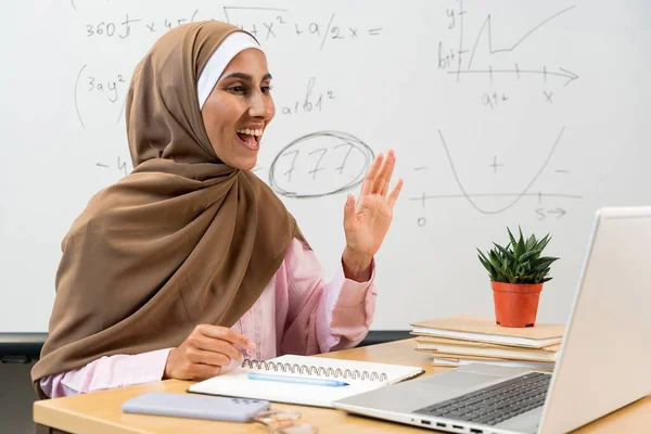 ヒジャブの若い東部の女性教師は ラップトップの前に机で座っている間 オンラインレッスン中に手を振って学生に挨拶します 教室で笑顔のイスラム教の先生 教育学習コンセプト — ストック写真