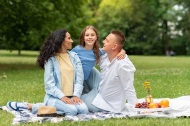 Karışık Irk Çifti ve Kızları Şehir Parkı 'nda bir gün tatil ve piknik yaptılar. Anne ve baba Sevgiyle Çocuklarına Bakar. Kız, anne ve babasına sarılıyor ve kameraya gülümsüyor..
