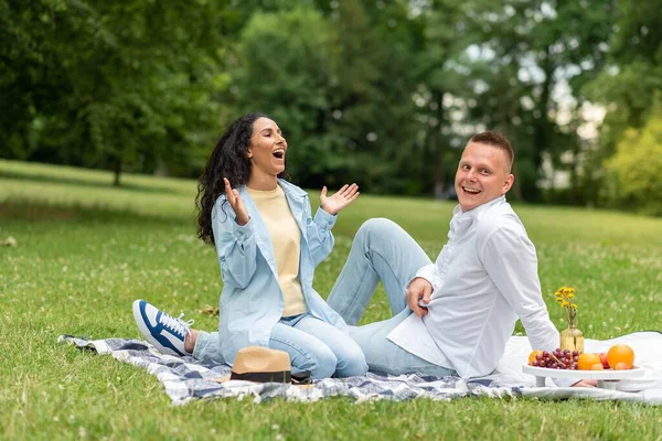 Funny joke. Pretty Latin girl, laughing and clapping her hands. Man an of Caucasian appearance sits next to her, smiling and laughing. Happy young couple in the park on a picnic spend leisure.