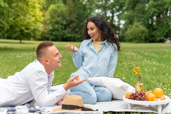 Couple of two lovely young people on a date in the park at a picnic. Latin girl gesticulates and laughs brightly. Caucasian man tells jokes story and amuses his partner. Fruit on a tray, a straw hat.