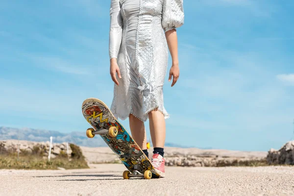 stock image A woman in an attractive silver dress keeps her foot on a skateboard. In the background is a beau-tiful blue sky.