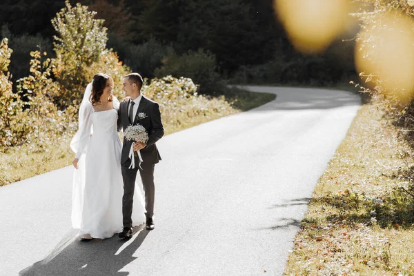 在他们的婚礼上 一对新婚夫妇一次不同寻常的 风景如画的散步 日落时分 一位身穿西服的年轻丈夫和一位穿着婚纱的美丽而纤细的新娘在草原上的路上 — 图库照片