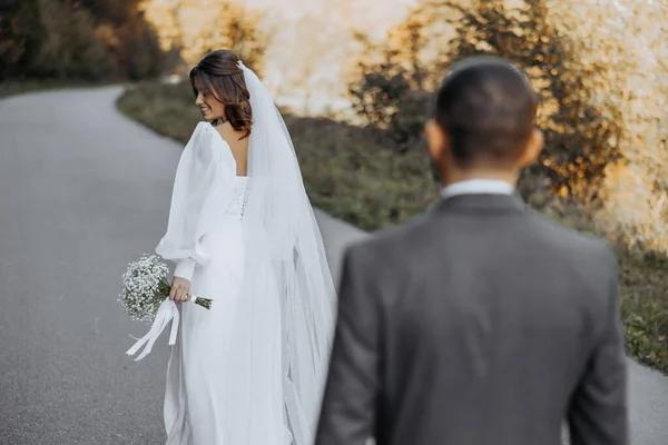新郎跟在新娘后面 沿着大路走着 新娘穿着一件轻盈漂亮的婚纱和一束吉普赛花 婚礼和婚纱照在天空下的概念 — 图库照片