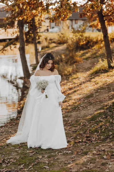 漂亮的新娘 穿着长长的婚纱 手里拿着美丽的花束 面带微笑 在树下的公园里美丽的夕阳下 — 图库照片