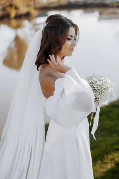 柔らかい光の中で湖の近くに長いベールとジプシーの花束を持つエレガントな白いドレスで美しい若い花嫁の肖像画 — ストック写真