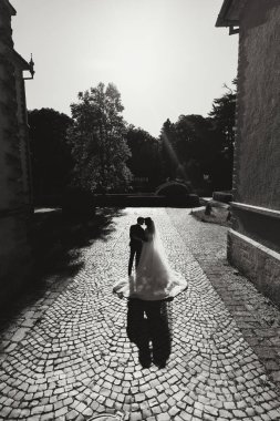 Gelin ve damat, gölgelerle batan güneşin ışığında dar bir sokakta dururlar. Siyah beyaz fotoğraf