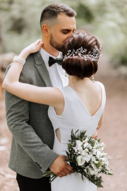 Damat bir buket çiçek tutuyor ve karısını kucaklıyor, tapınağını öpüyor. Gelinin arkası dönük. Fotoğraf ayrıntıları