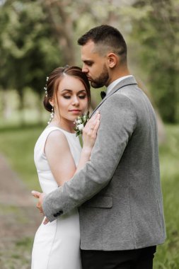 Gelin beyaz bir gece elbisesi içinde damat ile güzel bir saç stili ile poz verir, damat gelini nazikçe sarıp ona yaslanır. Portre fotoğrafı